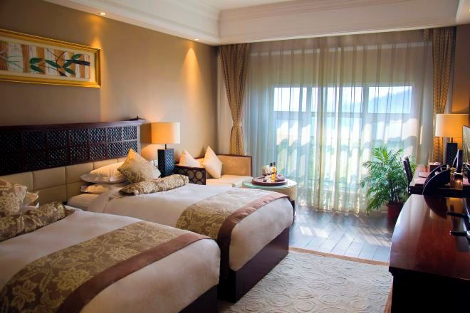 Quarto de hotel com duas camas, uma janela grande com cortinas, uma escrivaninha com luminária e uma planta em vaso.