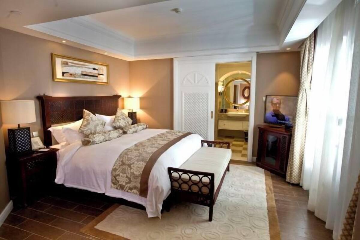 A bedroom with a large bed, bedside tables, a nightstand, a mirror, a television, and a bathroom visible through an archway.