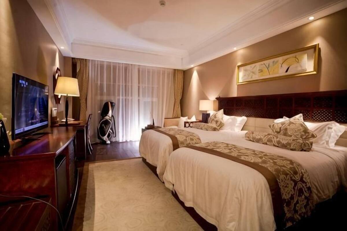 A hotel room with two beds, a TV, a desk, and a large window with curtains.