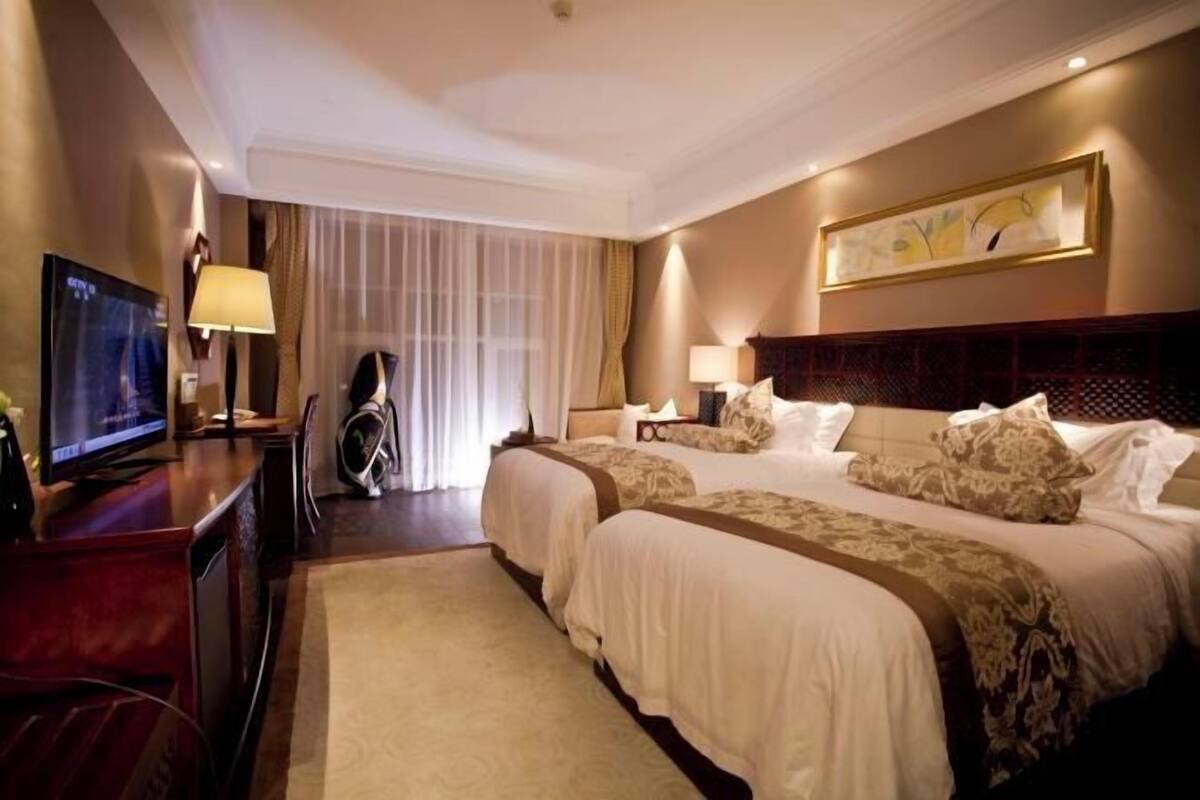 A hotel room with two beds, a TV, a desk, and a large window with curtains.