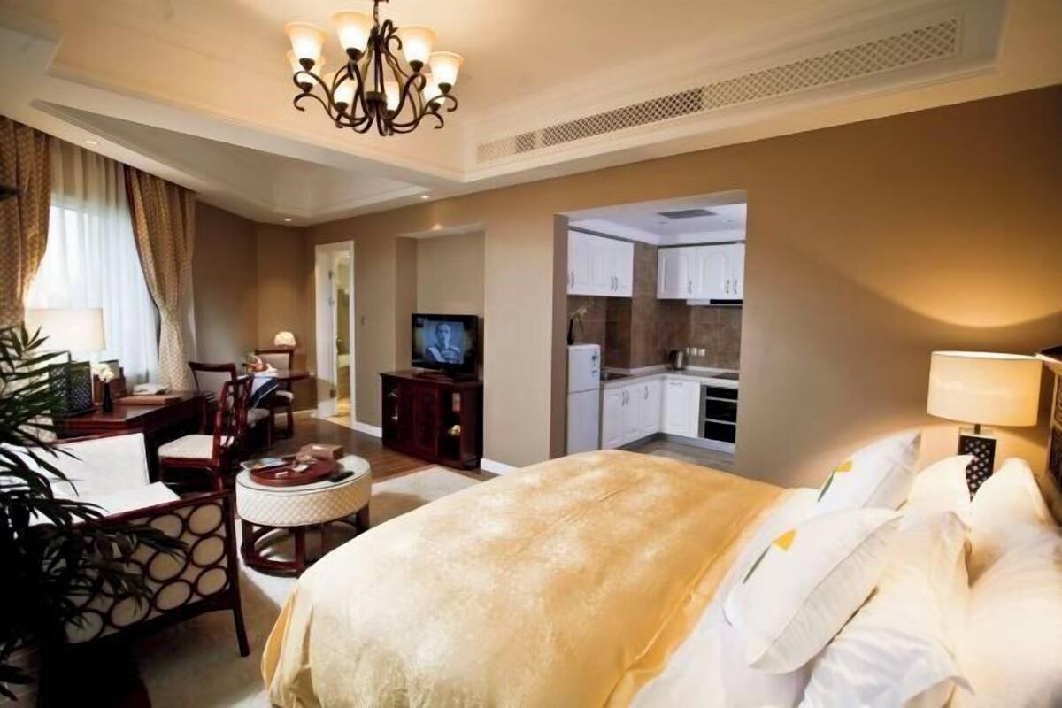 A hotel room with a bed, a TV, a desk with a chair, a small dining area, and a kitchenette.