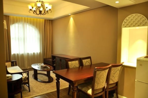 A hotel room with a dining area featuring a wooden table and chairs, a small round table, a chandelier, and a window with curtains. - Zhongcheng Holiday Hotel (Shijiazhuang)