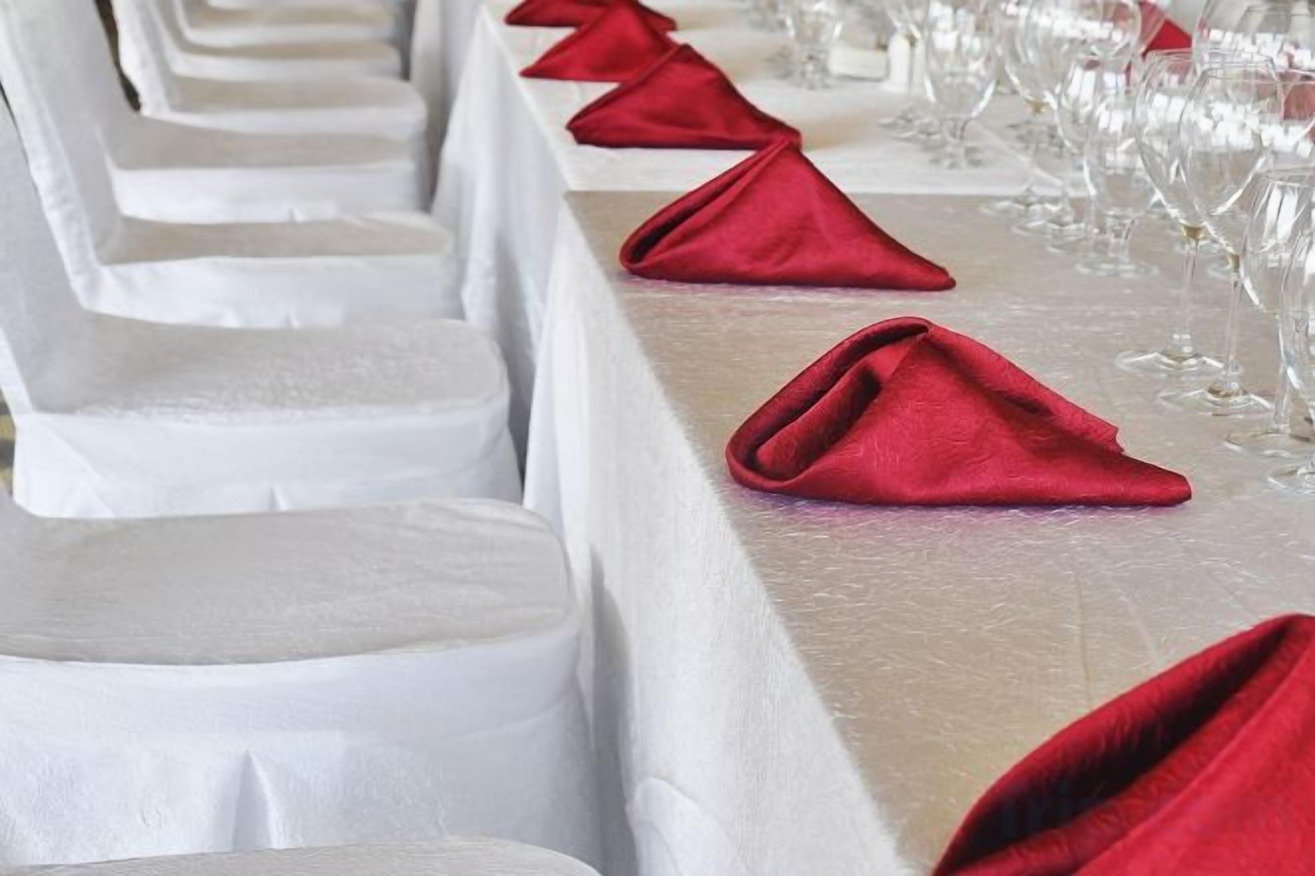 Fileira de cadeiras com capas brancas e guardanapos vermelhos sobre uma mesa, indicando um ambiente de jantar formal.