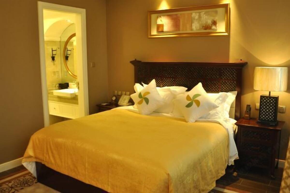 A hotel room with a bed, a nightstand, a lamp, a framed picture, and a bathroom visible through an open door.