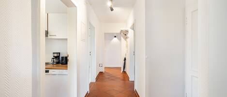 A hallway with white walls, a tiled floor, and a kitchen area visible on the left.