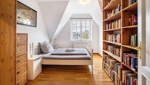 Un dormitorio con ventanal elevado, cama, mesita de noche, estantería y armario de madera.