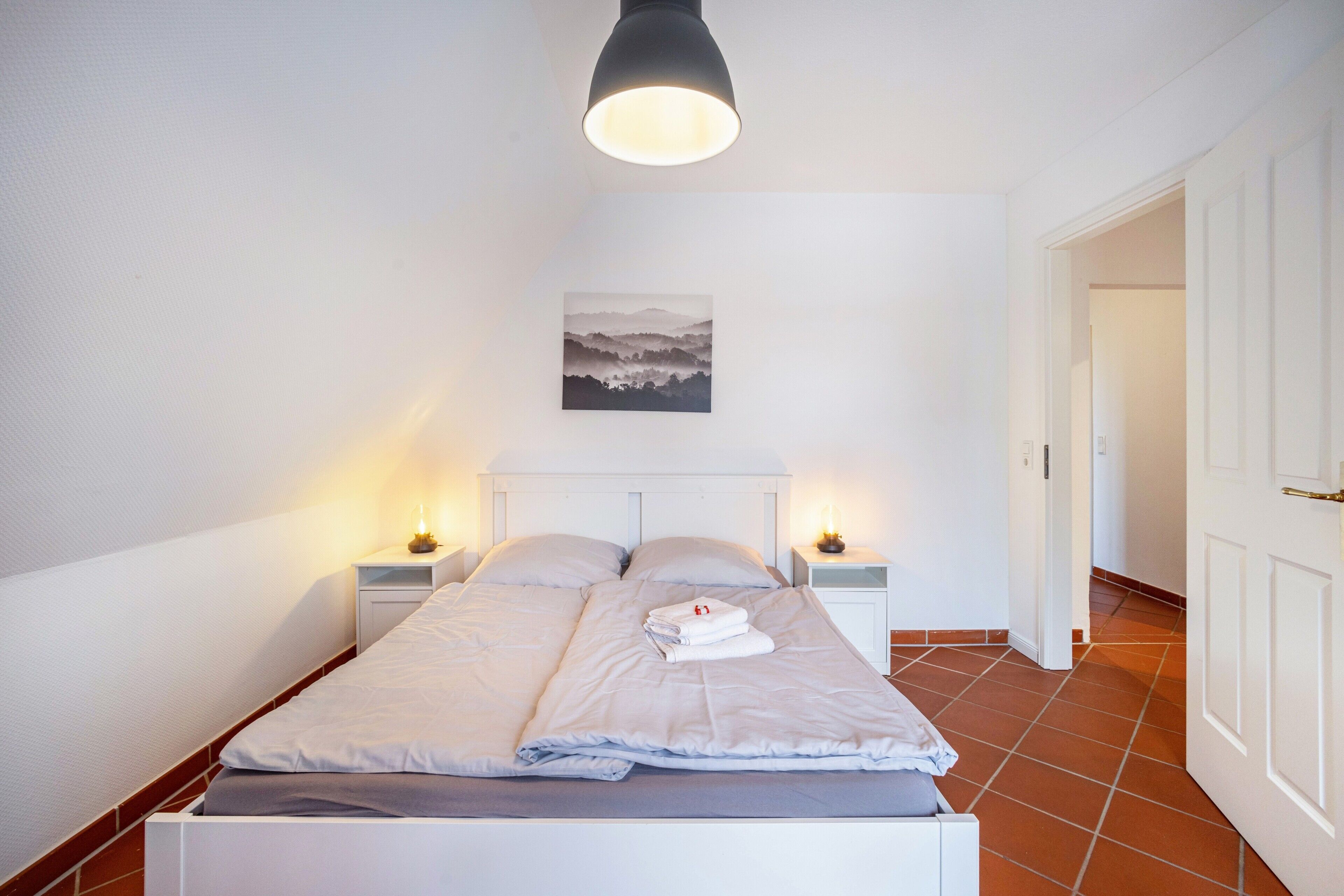 Een keurig opgemaakt bed met een wit hoofdbord, twee nachtkastjes met lampen, een aan de muur hangend schilderij, en een deur aan de rechterkant.