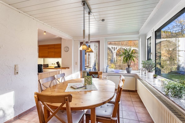Un espace repas comprenant une table et des chaises en bois, un luminaire suspendu et une vue sur l’extérieur grâce à une grande fenêtre.