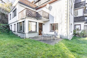 Une maison à deux étages avec un balcon, une terrasse avec une table et des chaises, et un jardin.