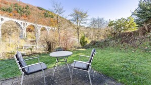 A patio with two chairs and a table, a stone bridge, and a hillside with trees. - Haus Sonneck - Apartment 5 with Mountain View, Private Terrace, and Wi-Fi (Willingen (Upland))