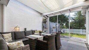 Ein überdachter Patio-Bereich mit einer Couch, Korbstühlen, einem Tisch und Blick auf den Garten.