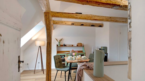 Un espacio tipo loft con vigas de madera expuestas, una mesa de comedor con sillas, una lámpara y una zona de cocina al fondo.