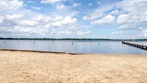 Een strand met een houten steiger die zich uitstrekt over kalm water, een zandstrand en een verre boomlijn onder een licht bewolkte lucht.