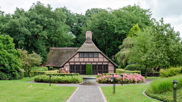 Een traditioneel, Tudor-stijl gebouw met rieten dak, omgeven door een verzorgde tuin met een geplaveid pad naar de ingang.