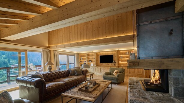 A spacious living room with a leather sofa, a stone fireplace, and a television mounted on a wooden wall. - Apartment 'Steinbock B' with Mountain View, Balcony and Wi-Fi (Klosters-Serneus)