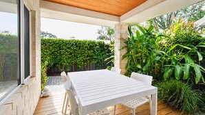 Une terrasse couverte avec une table et des chaises blanches, entourée de verdure et d’un mur de briques.