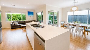 Une cuisine moderne équipée d’un lave-vaisselle, un coin repas avec une table et des chaises, un canapé et de grandes fenêtres offrant une vue sur la verdure.