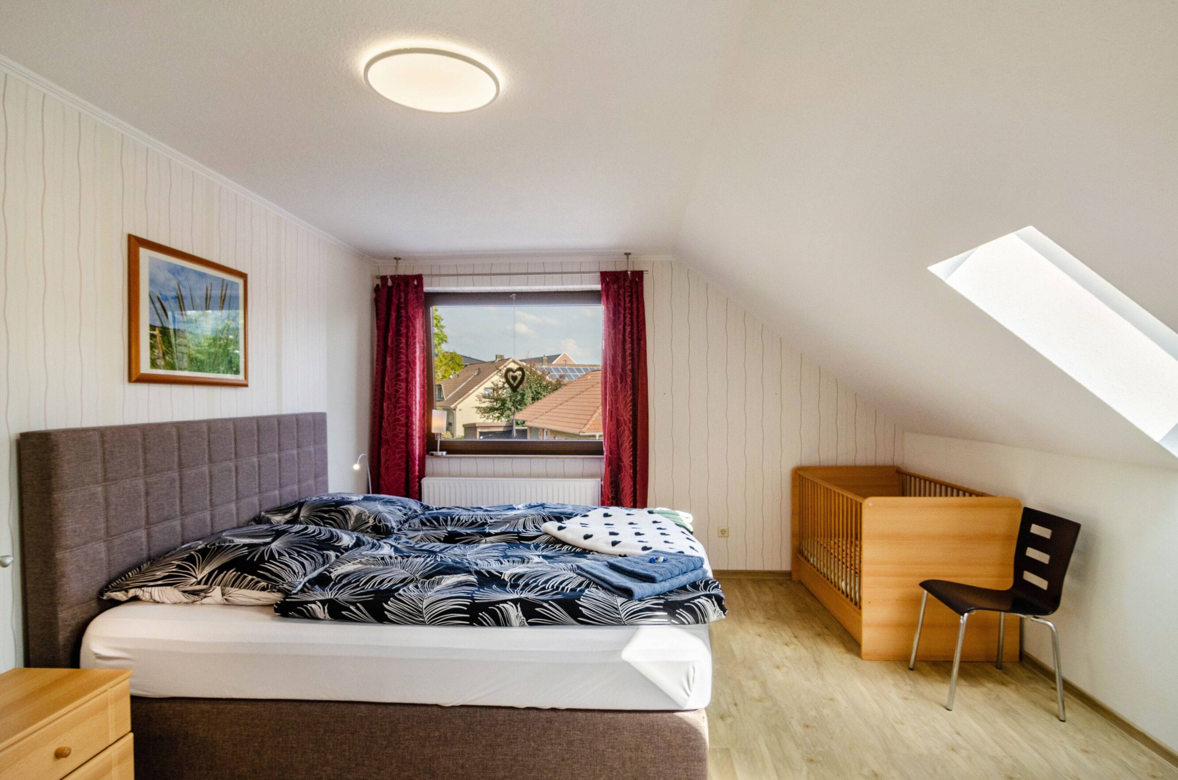 Een slaapkamer met een bed, een ledikant, een stoel, een raam met gordijnen, een foto aan de muur en een dakraam.