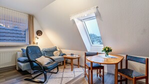 A loft bedroom with a sloped ceiling, a sofa, a rocking chair, a dining table with chairs, and a window with a view.