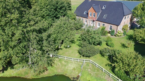 A large house with a sloped roof, surrounded by trees and a small pond.