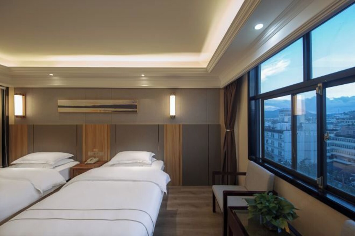 Habitación de hotel con dos camas, un ventanal amplio con vistas a la ciudad, una mesita de noche con lámpara y aire acondicionado instalado en la pared.