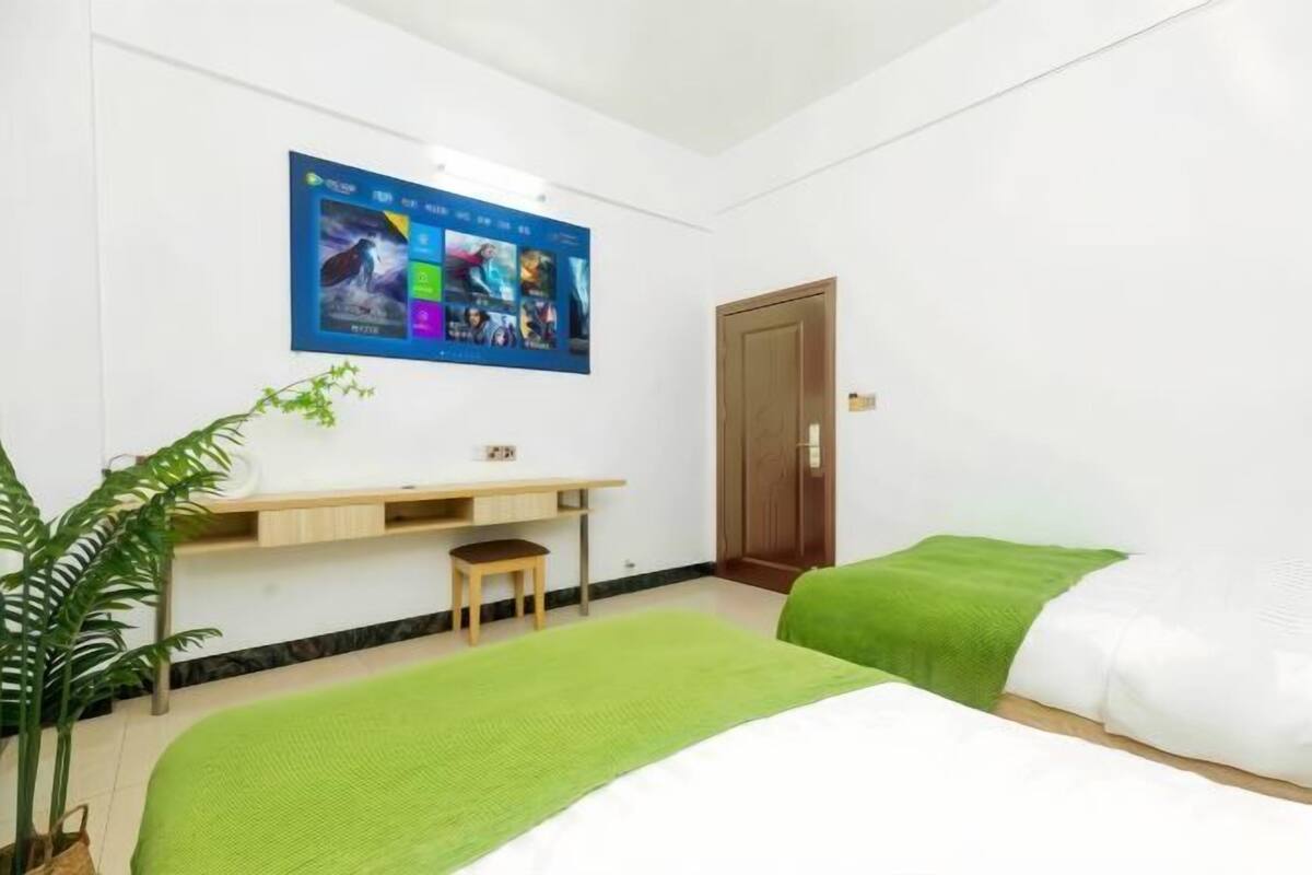 A hotel room with a bed, a desk, a chair, a potted plant, and a wall-mounted TV displaying various channels.