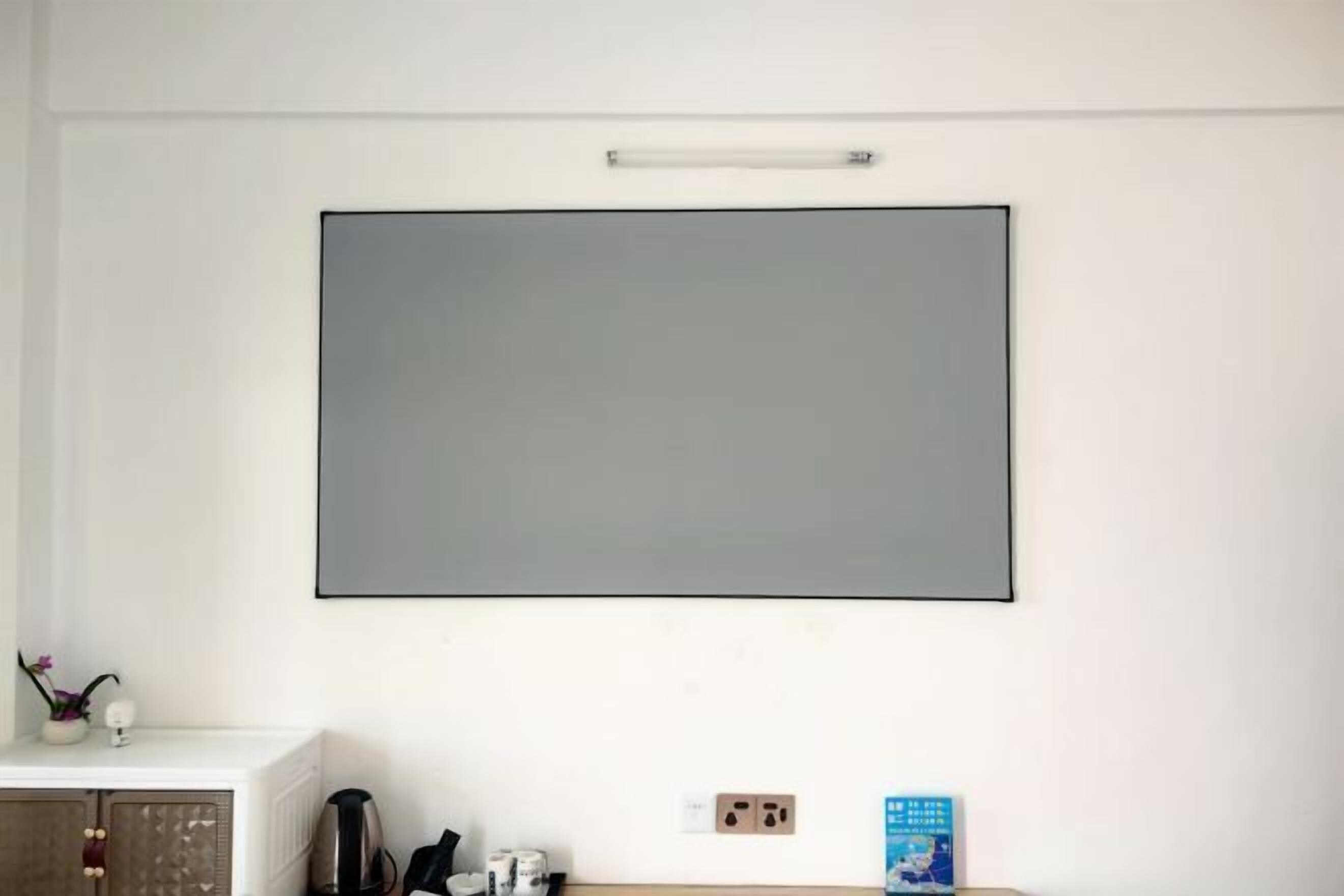 A room with a large, blank wall-mounted TV, a white cabinet with a decorative item, and a small table with a book and a kettle.