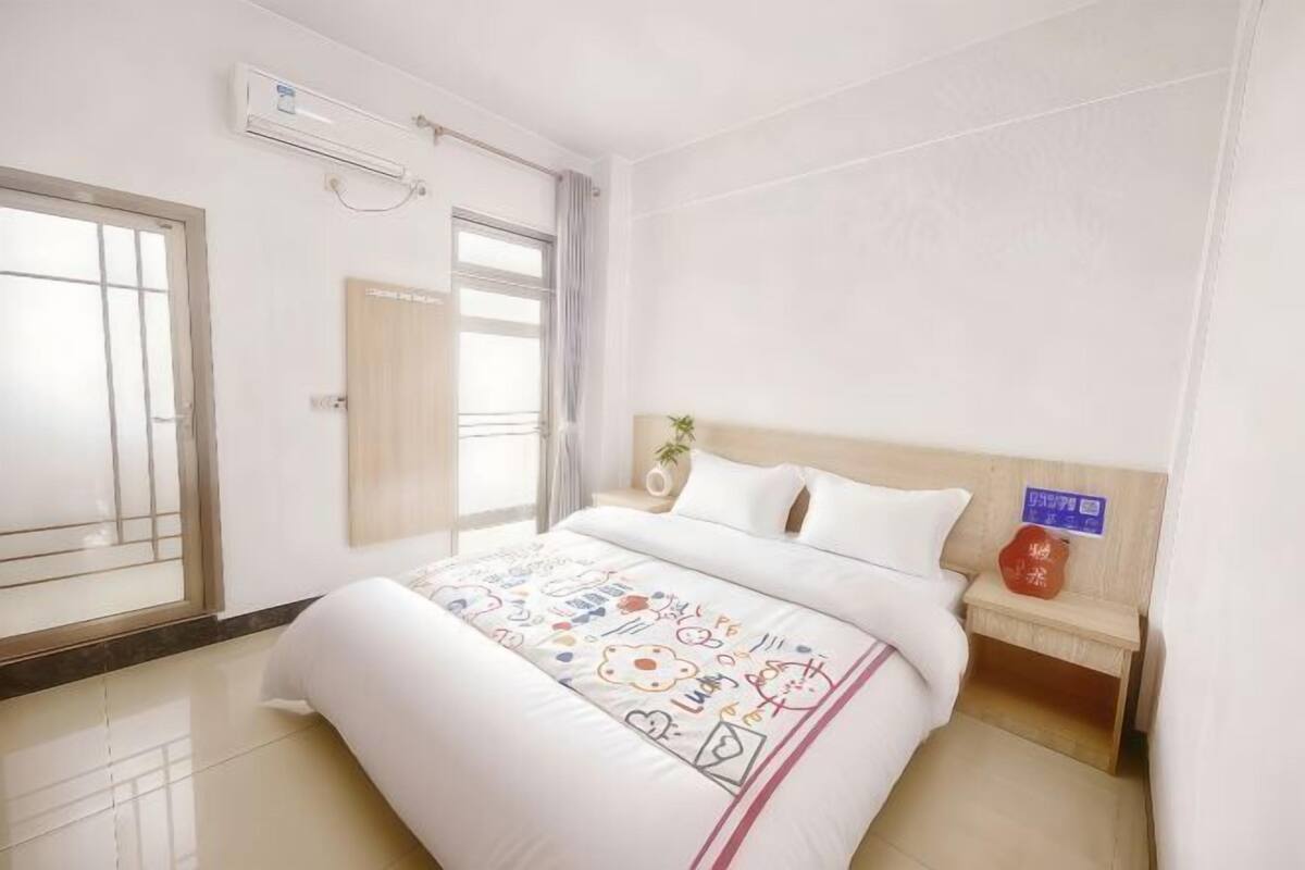 A hotel room with a bed, a bedside table, a wall-mounted air conditioner, and a window with curtains.