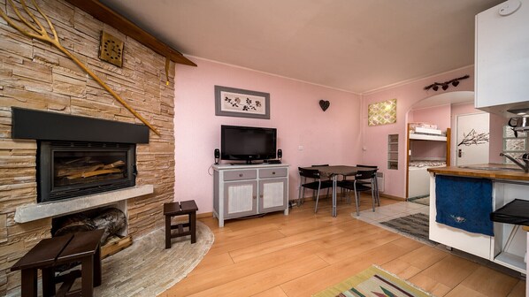 A cozy living room with a stone fireplace, a television, a dining area, and a kitchen with a sink and counter.