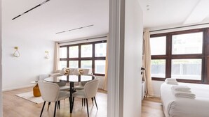 Moderni hotellihuone, jossa on pyöreä ruokapöytä, sohva, siististi taitetuin pyyhkein varustettu sänky ja suuret ikkunat, joista on näkymä ulos.