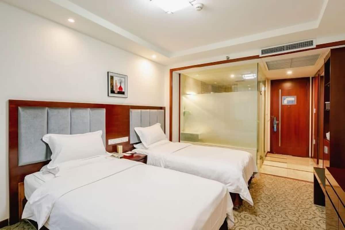 A hotel room with two beds, a wooden headboard, a small bedside table, a nightstand, a framed picture on the wall, and a sliding door leading to a bathroom.