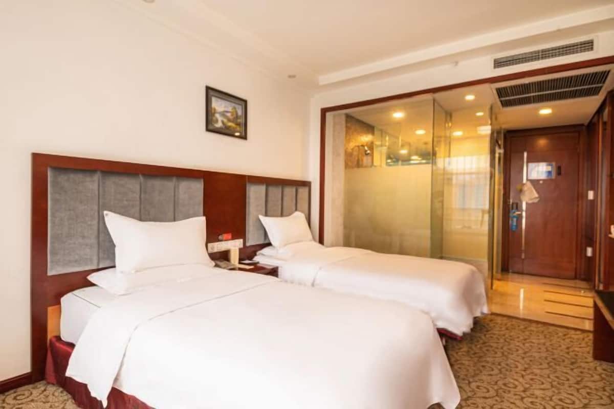 A hotel room with two beds, a wooden headboard, a bedside table, a nightstand, a framed picture on the wall, and a glass door leading to a bathroom.