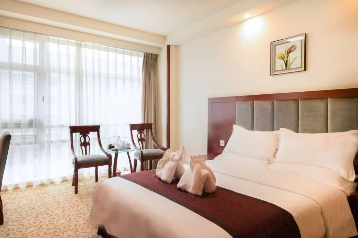 A hotel room with a bed, two chairs, a small table, and a window with sheer curtains.