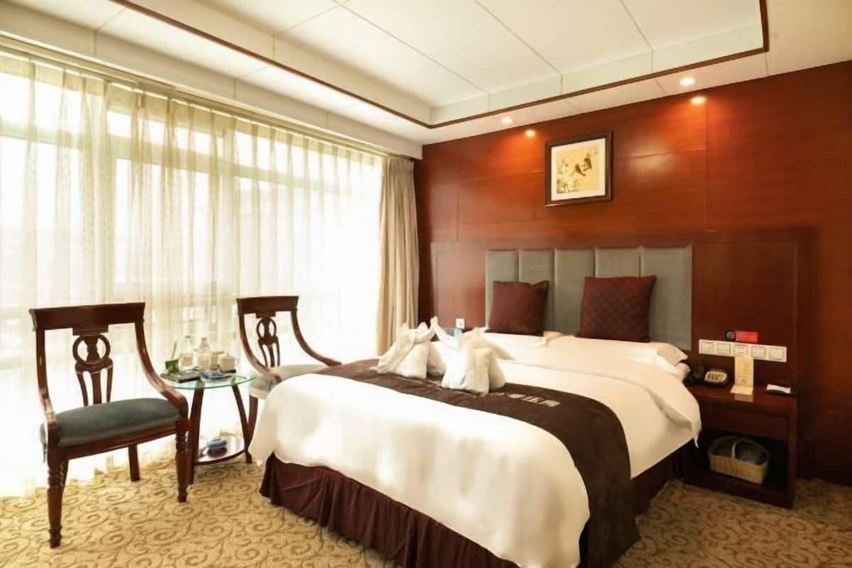 A hotel room with a large bed, a small table with a chair, a nightstand, and a window with sheer curtains.