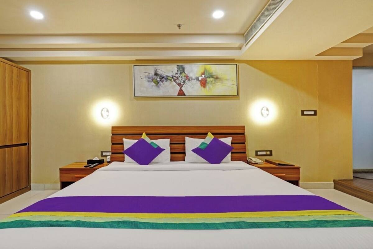 A hotel room with a large bed, wooden headboard, bedside tables, a nightstand, a wardrobe, and a painting on the wall.