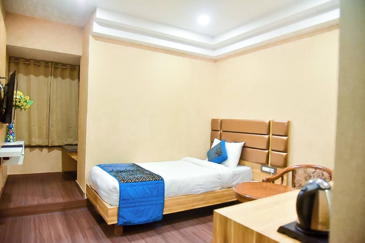 A hotel room with a single bed, a wooden headboard, a bedside table, a chair, and a small desk with a kettle.