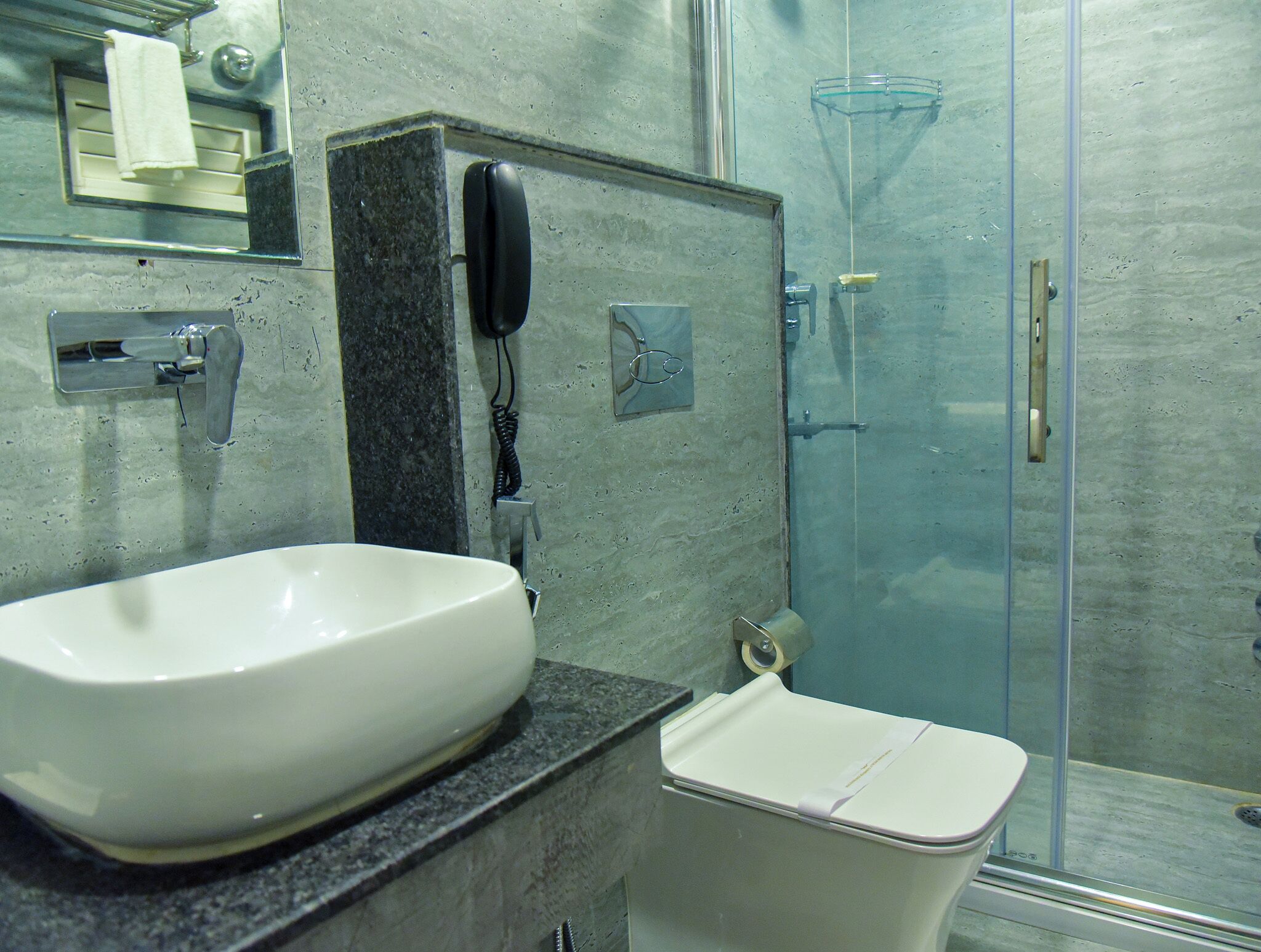 A bathroom with a white sink, a toilet, a glass shower enclosure, and a telephone mounted on the wall.