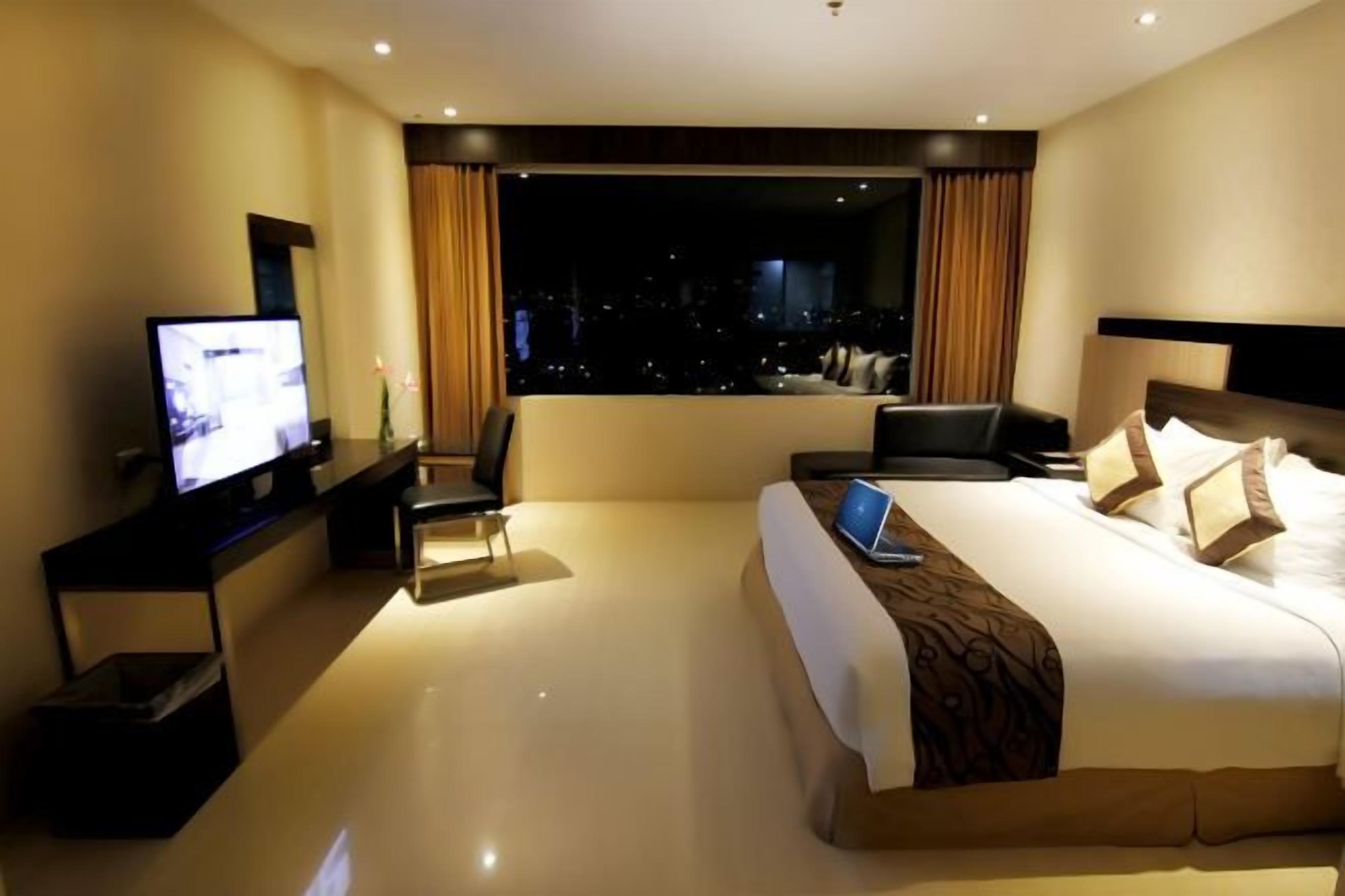 A hotel room with a large bed, a desk with a laptop, a television, and a view of the city at night.