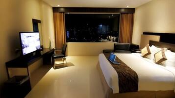 A hotel room with a large bed, a desk with a laptop, a television, and a view of the city at night.