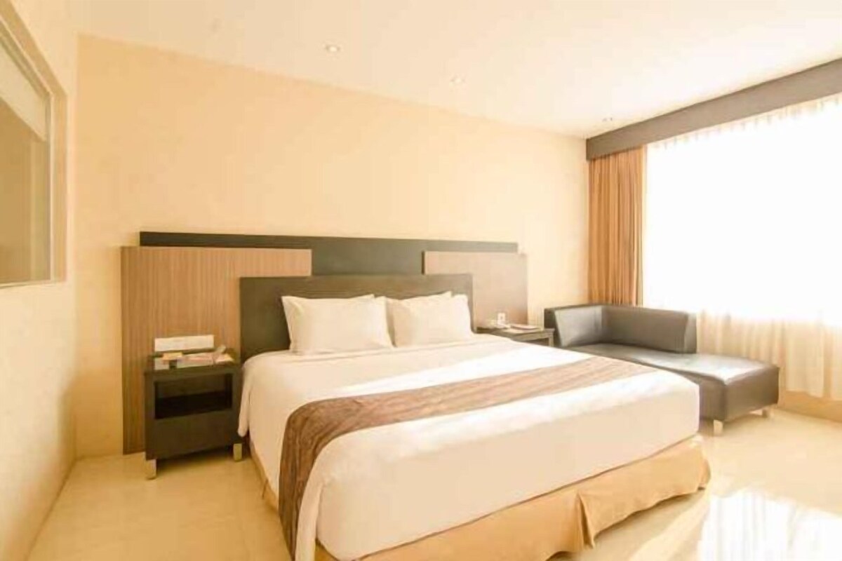 Ein Hotelzimmer mit Bett, Nachttischen, einem Schreibtisch und einem mit Vorhängen versehenen Fenster.