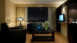 A modern hotel room with a large window offering a city view, a black leather sofa, a glass coffee table, and a flat-screen TV.