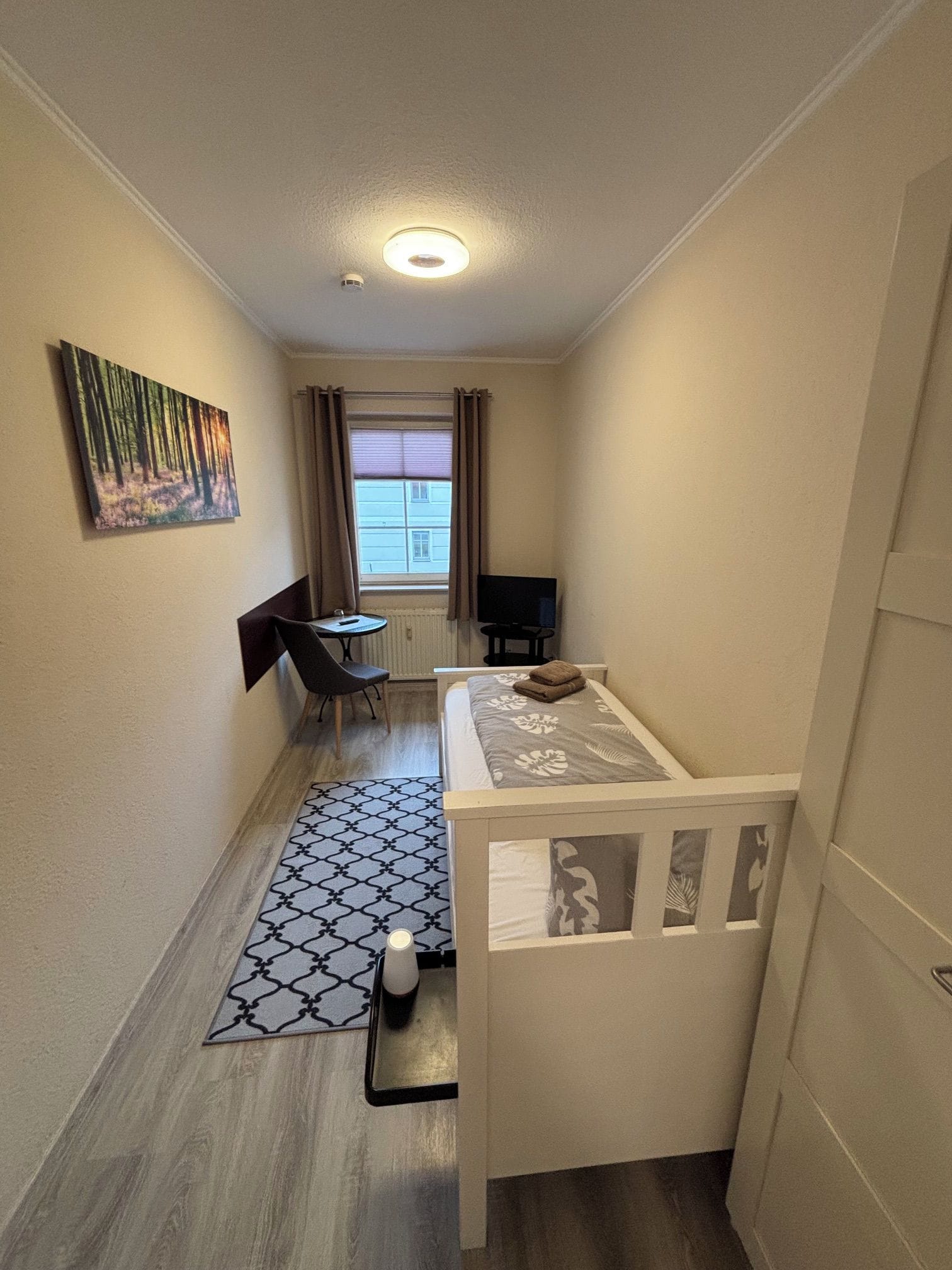 A narrow hallway with a bed, a desk, a chair, a TV, and a window with curtains.