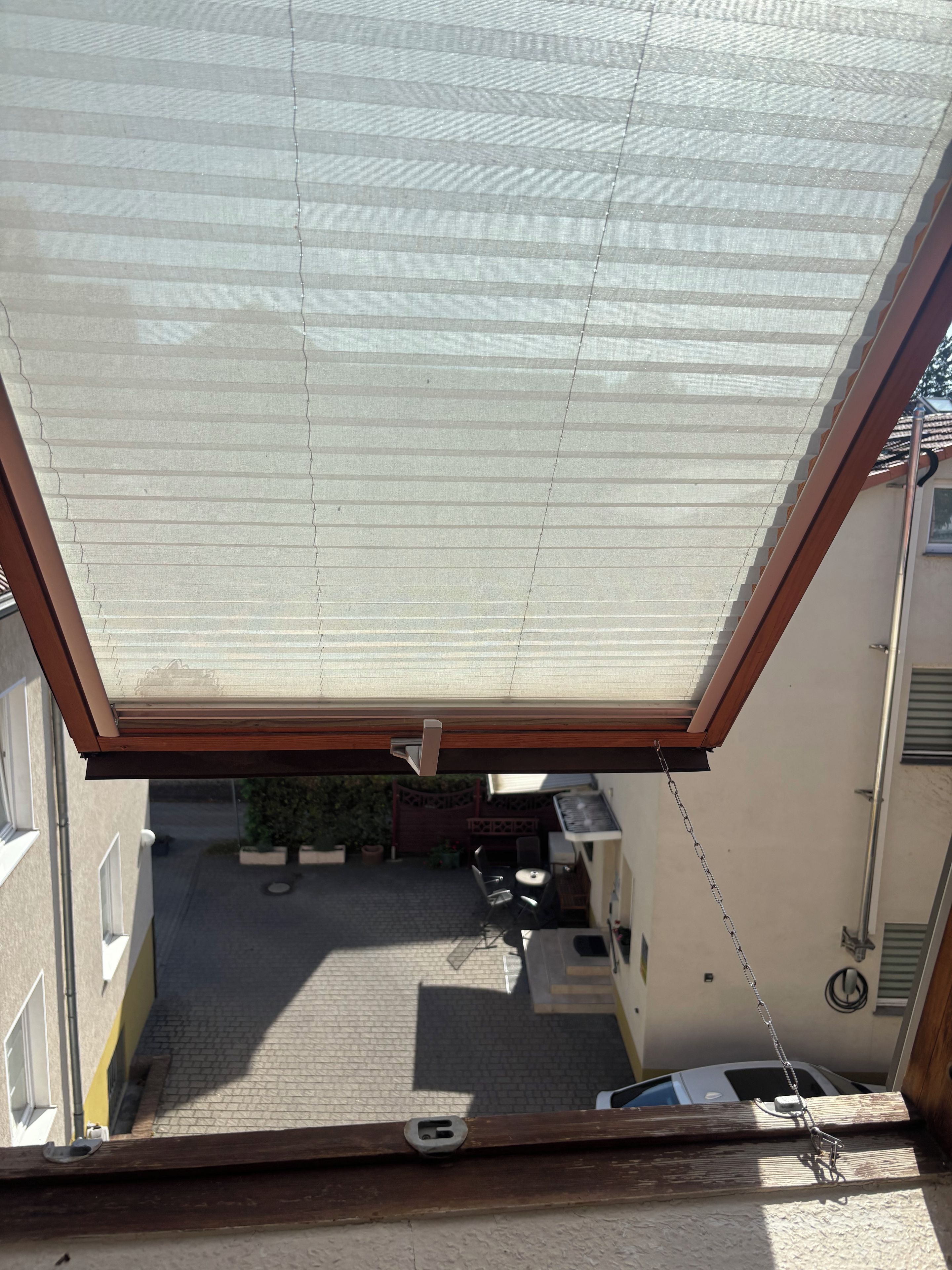 Blick durch ein Dachfenster auf einen Innenhof mit einem geparkten Auto, einem Tisch und Stühlen.