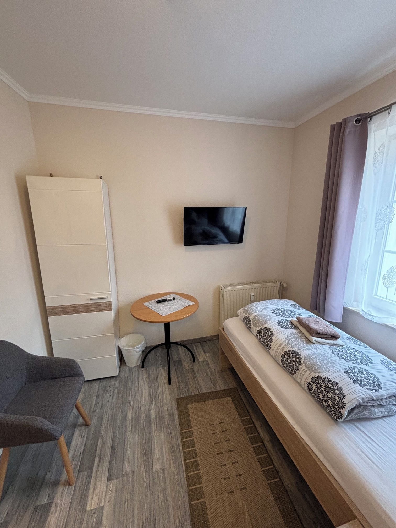 A small, neatly arranged room with a bed, a chair, a small table, a television, and a radiator.