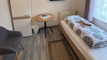 A small, neatly arranged room with a bed, a chair, a small table, a television, and a radiator.