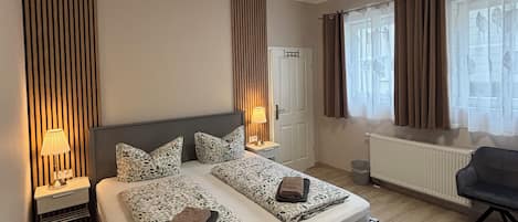 A modern hotel room with a bed, bedside tables, a bench, a radiator, and a window with curtains.