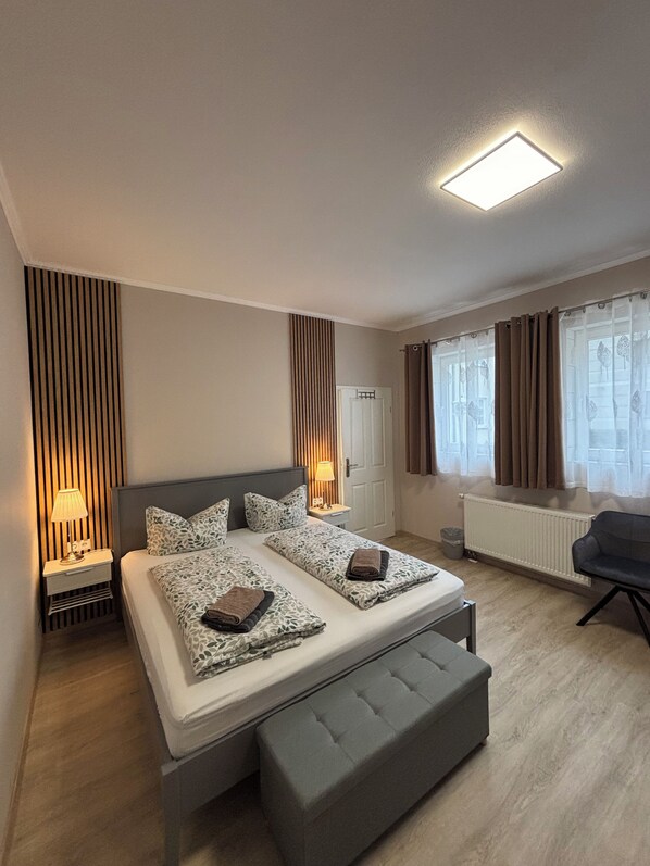Una habitación de hotel moderna con cama, mesitas de noche, un banco, un radiador y una ventana con cortinas.