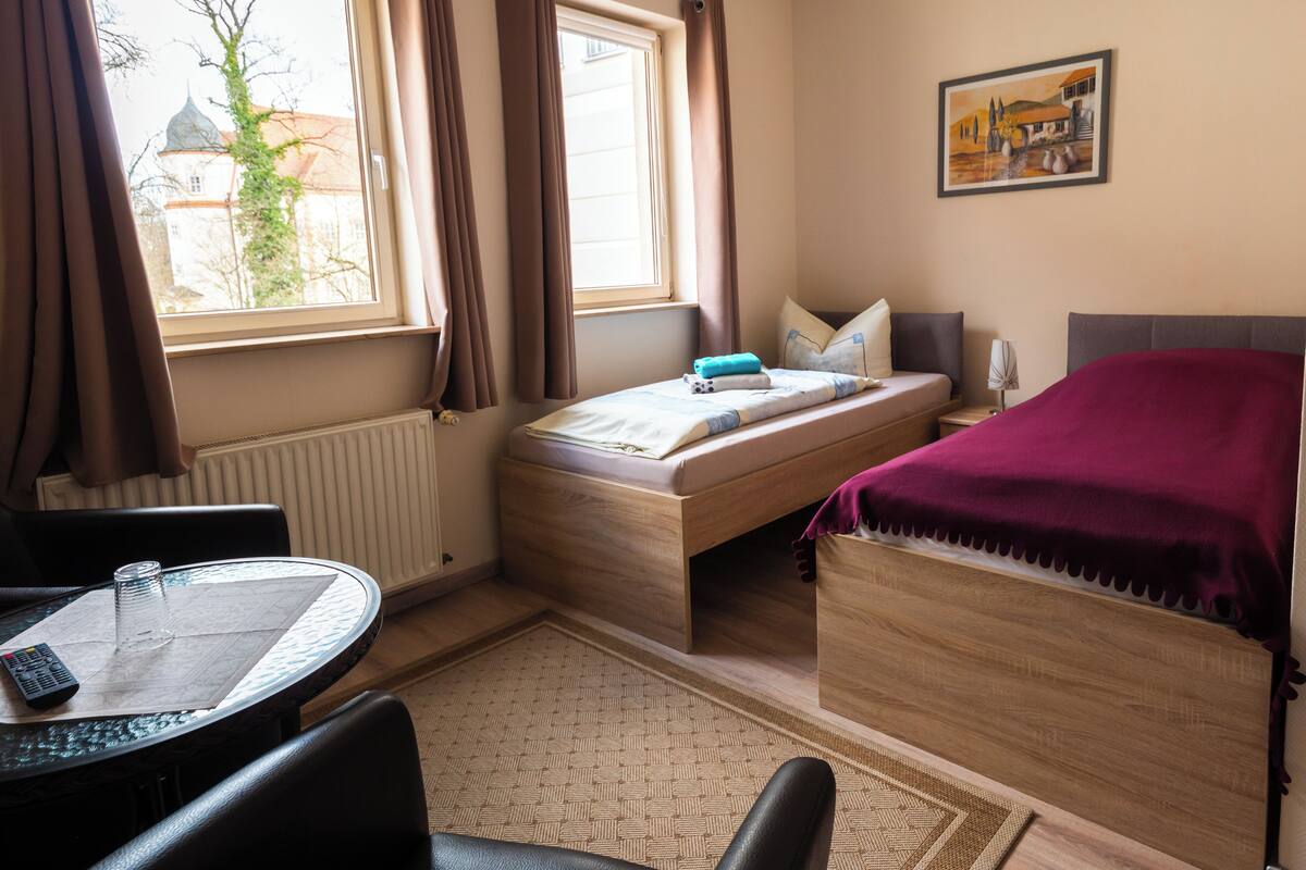 A compact room with a bed, a small table with a remote control, a window with curtains, and a painting on the wall.