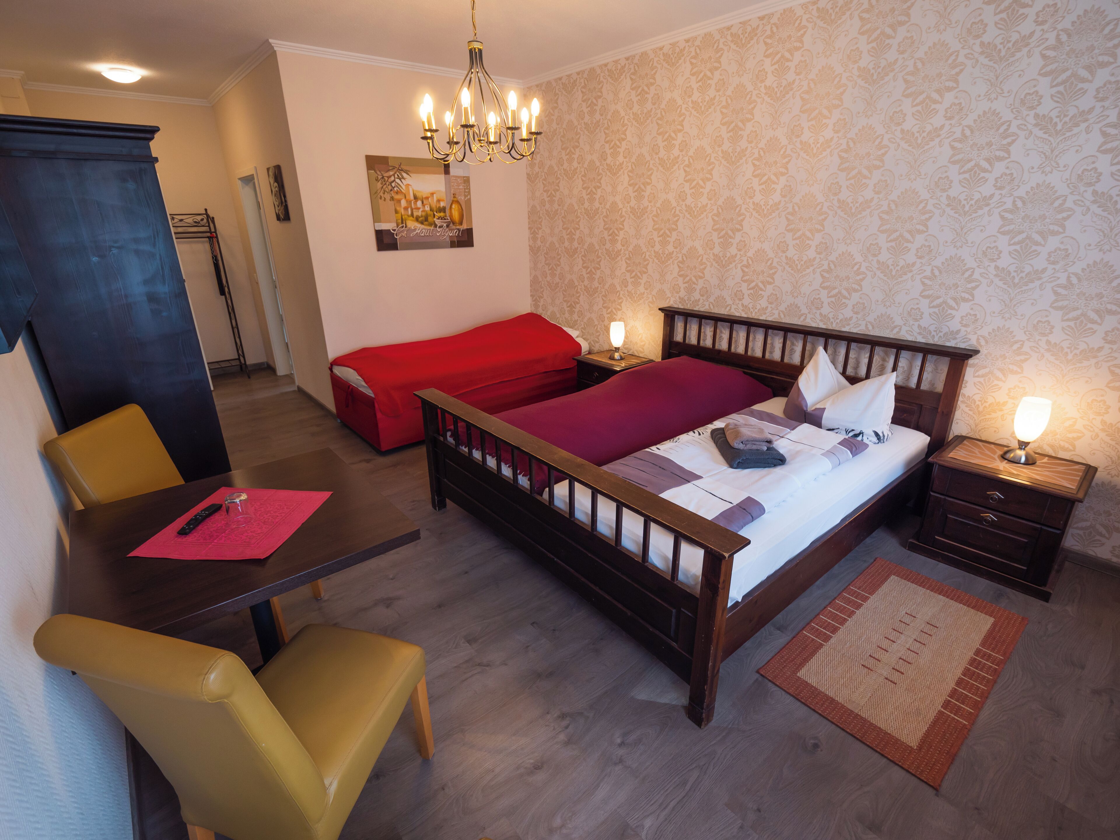 A bedroom with a wooden bed, a red sofa, a dining table with chairs, a chandelier, and a nightstand with a lamp.
