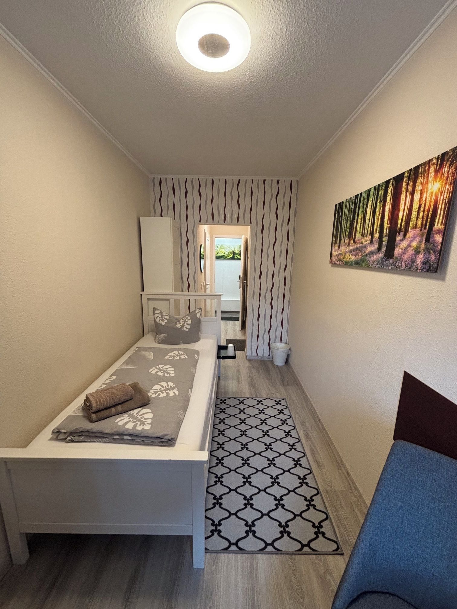 A bedroom with a bed, a chair, a painting, a patterned rug, and a wardrobe.